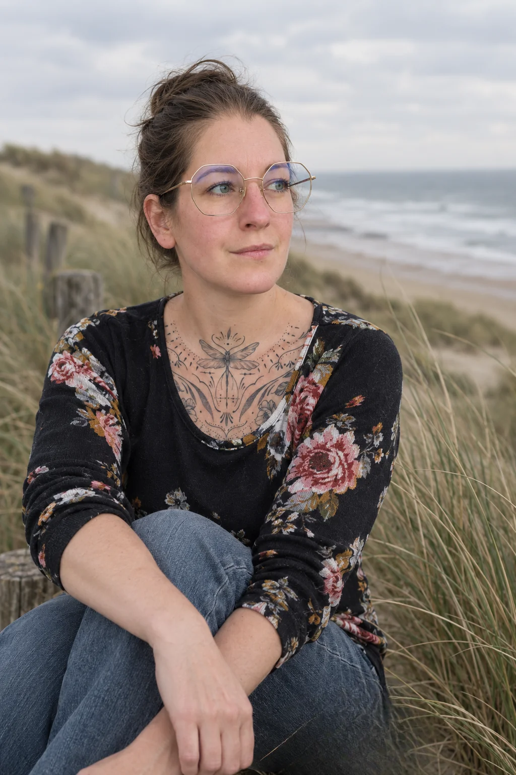 Miriam van Brouwershaven van De Libelle Shift zit in een gebloemde trui en met bril in de duinen met de zee op de achtergrond